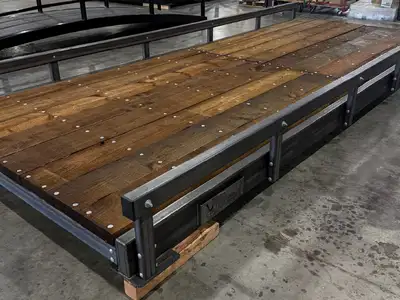 The deck of a raw steel bridge in a workshop, dark stained timber boards bolted down between steel bump rails, with an arched truss standing behind it.