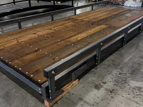 The deck of a raw steel bridge in a workshop, dark stained timber boards bolted down between steel bump rails, with an arched truss standing behind it.