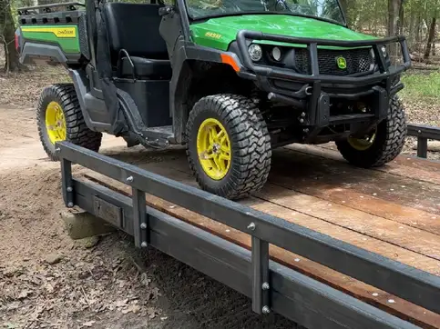A green utility vehicle parked on a raw steel bridge with timber decking and a low bump rail, on a dirt track in open woodland.