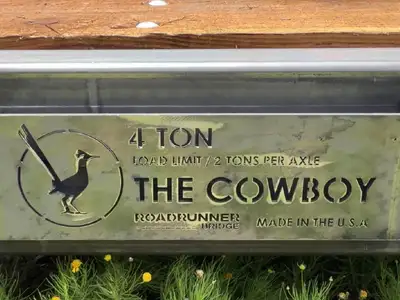 A cast steel plate on the side of a bridge reading four ton, load limit two tons per axle, The Cowboy, Roadrunner Bridge, Made in the U.S.A., with a roadrunner mark beside the text, weathered to a grey green, timber decking above and grass below.