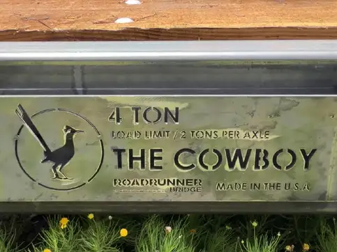 A cast steel plate on the side of a bridge reading four ton, load limit two tons per axle, The Cowboy, Roadrunner Bridge, Made in the U.S.A., with a roadrunner mark beside the text, weathered to a grey green, timber decking above and grass below.