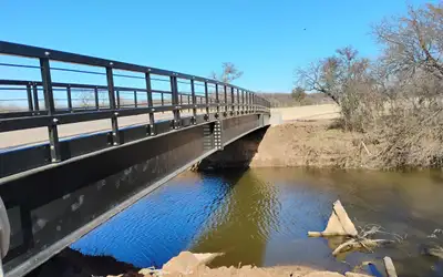 A long black bridge with a horizontal railing seen along its length, crossing a creek between poured concrete abutments in winter scrub.