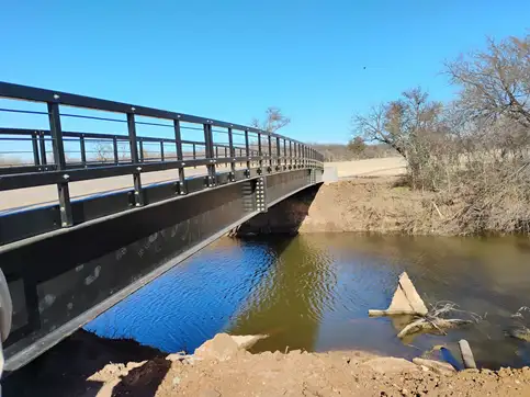 A long black bridge with a horizontal railing seen along its length, crossing a creek between poured concrete abutments in winter scrub.