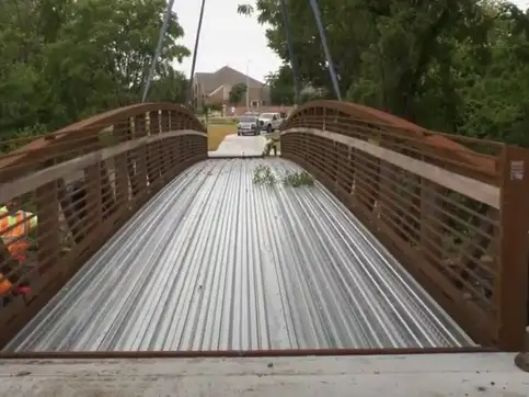 Two workers in hard hats and high visibility vests standing either side of a finished bridge deck, looking along it toward a road and houses.