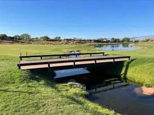A black steel bridge with a timber deck and a low rail along each side, crossing a narrow creek on a golf course, with a pond, red earth and desert houses under a clear blue sky behind.