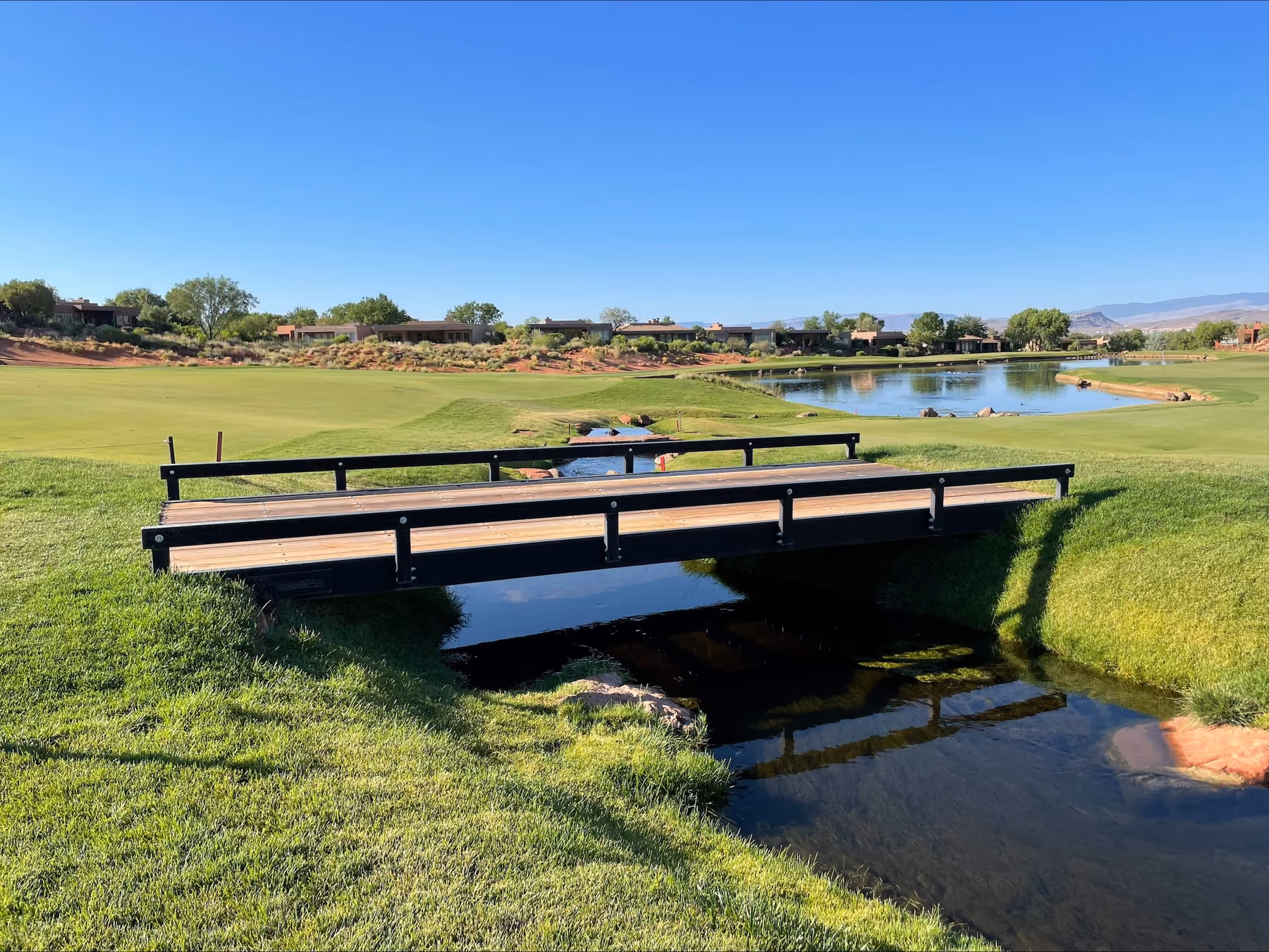 A black steel bridge with a timber deck and a low rail along each side, crossing a narrow creek on a golf course, with a pond, red earth and desert houses under a clear blue sky behind.
