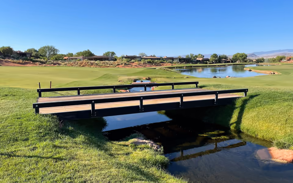 A black steel bridge with a timber deck and a low rail along each side, crossing a narrow creek on a golf course, with a pond, red earth and desert houses under a clear blue sky behind.