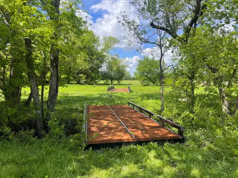 A timber-decked steel bridge with a low side rail crossing a wooded creek into a sunlit meadow, with a second bridge visible in the grass beyond.