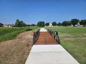 A short black steel truss bridge with a stained timber deck carrying a concrete path across a grassy drainage swale in a park, under a clear sky.