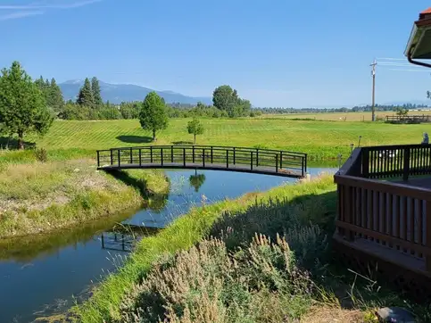 A slender steel truss bridge spanning a still creek in a green pasture, with a timber deck rail in the near foreground and mountains along the horizon.