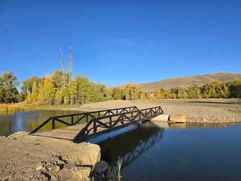 A black steel truss bridge crossing a still pond onto a concrete abutment, with yellow autumn cottonwoods and dry hills behind.