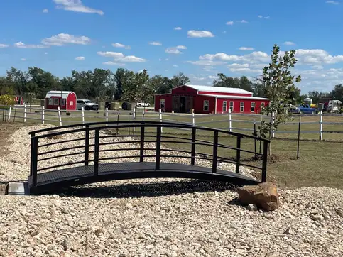 A black arched steel bridge with horizontal railings set on a bed of pale river rock, with red barns and a white rail fence behind it.