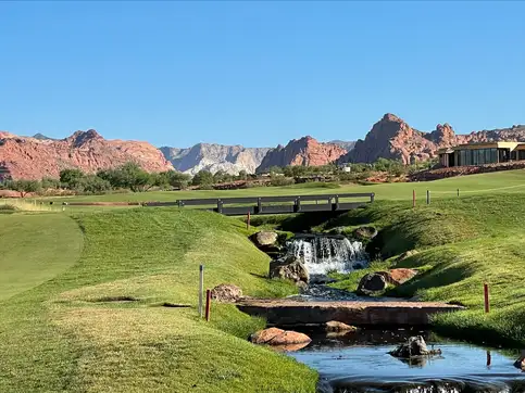 A steel bridge with a horizontal railing carrying a path over a rocky stream and small waterfall on a golf course, with red sandstone cliffs on the skyline behind.