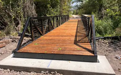 A black steel truss bridge with horizontal bars between its diagonals and a stained timber deck, seated on a new concrete footing at the edge of dense woodland.