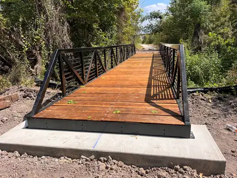 A black steel truss bridge with horizontal bars between its diagonals and a stained timber deck, seated on a new concrete footing at the edge of dense woodland.