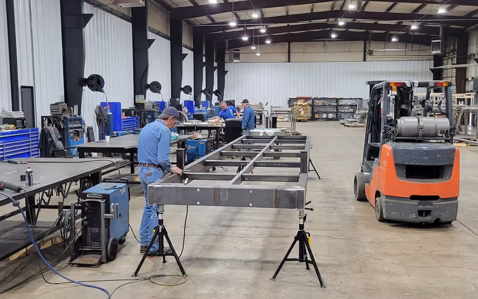 A steel bridge frame on stands in a fabrication shop, with welders working at benches and a forklift alongside.