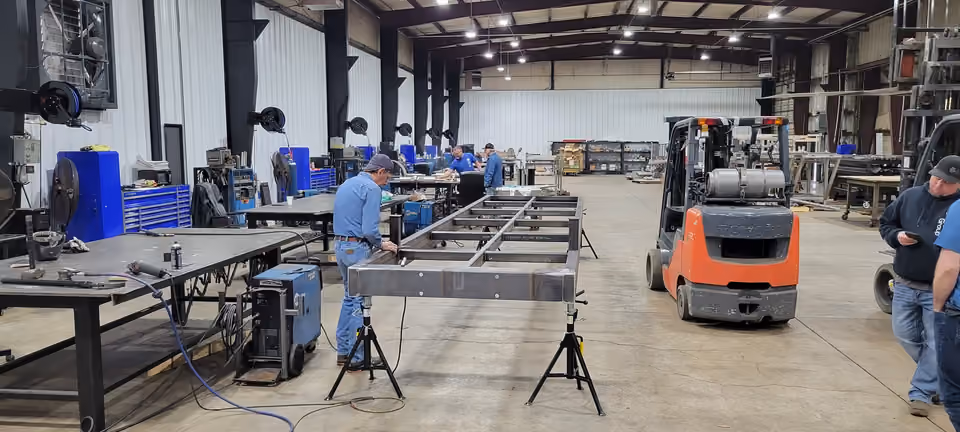 A steel bridge frame on stands in a fabrication shop, with welders working at benches and a forklift alongside.