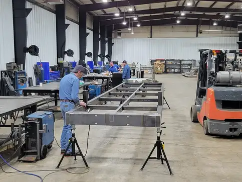A steel bridge frame on stands in a fabrication shop, with welders working at benches and a forklift alongside.