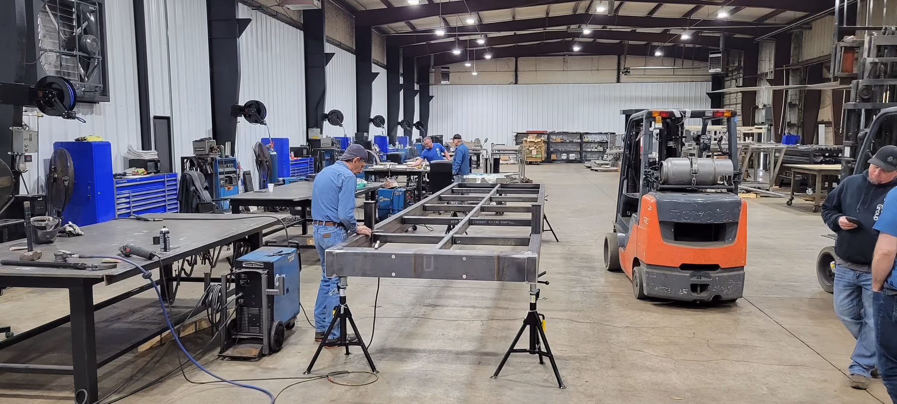 A steel bridge frame on stands in a fabrication shop, with welders working at benches and a forklift alongside.