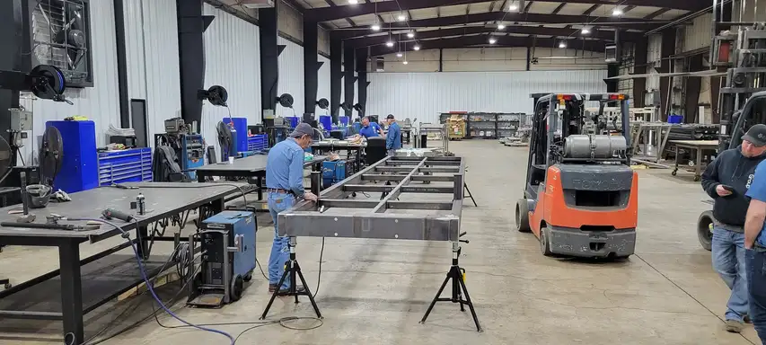 A steel bridge frame on stands in a fabrication shop, with welders working at benches and a forklift alongside.