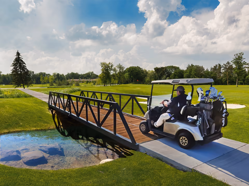A golf cart carrying two bags of clubs crossing a black steel truss bridge with timber decking over a pond on a golf course.