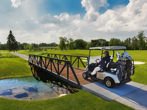 A golf cart carrying two bags of clubs crossing a black steel truss bridge with timber decking over a pond on a golf course.