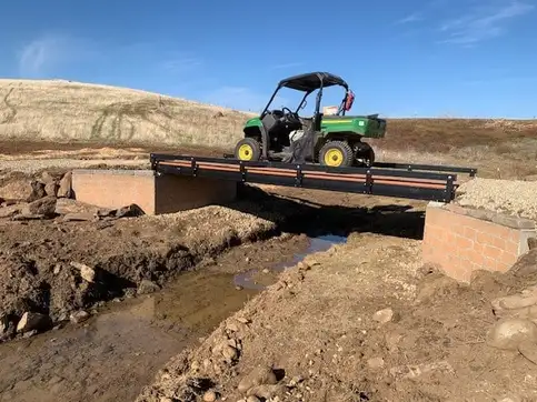 A green utility vehicle crossing a flat bolt-together bridge seated on block abutments over a freshly cut channel, with bare earth either side and dry hills behind.