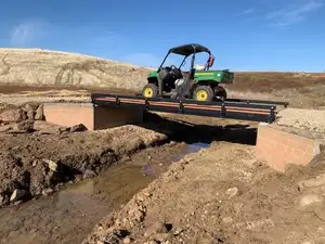A green utility vehicle crossing a flat bolt-together bridge seated on block abutments over a freshly cut channel, with bare earth either side and dry hills behind.