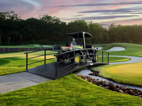 A course maintenance vehicle with a driver crossing a flat steel bridge with a horizontal railing over a stream on a golf course, under a pink and grey evening sky.