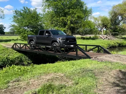 A black pickup truck stopped partway across a steel truss bridge over a grassed drainage channel, in open pasture with trees behind.