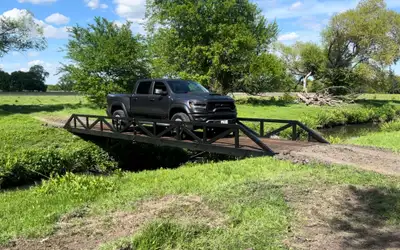 A black pickup truck stopped partway across a steel truss bridge over a grassed drainage channel, in open pasture with trees behind.