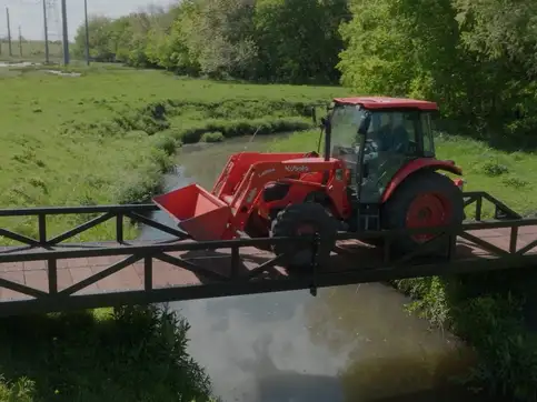 A red tractor with a front loader crossing a black steel truss bridge over a still pond, seen from above, in a green pasture.