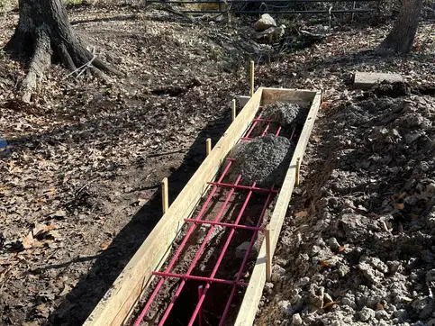 Timber formwork and pink rebar set in an excavated trench for a bridge abutment, in bare winter woodland.