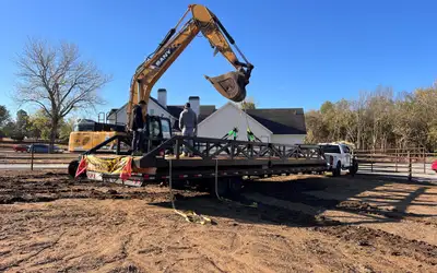 A steel truss bridge strapped to a gooseneck trailer, rigged with slings to an excavator, with two crew members standing on the deck.