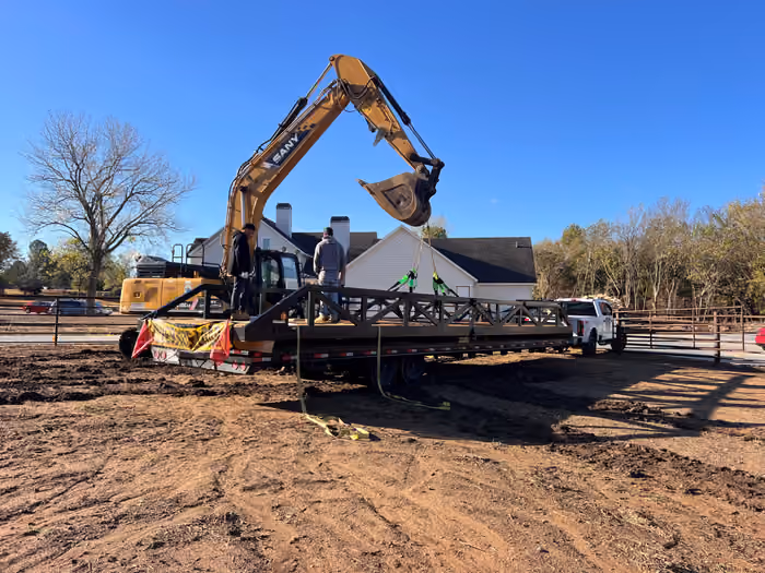 A steel truss bridge strapped to a gooseneck trailer, rigged with slings to an excavator, with two crew members standing on the deck.