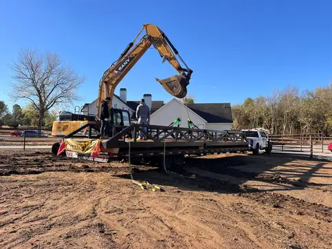 A steel truss bridge strapped to a gooseneck trailer, rigged with slings to an excavator, with two crew members standing on the deck.