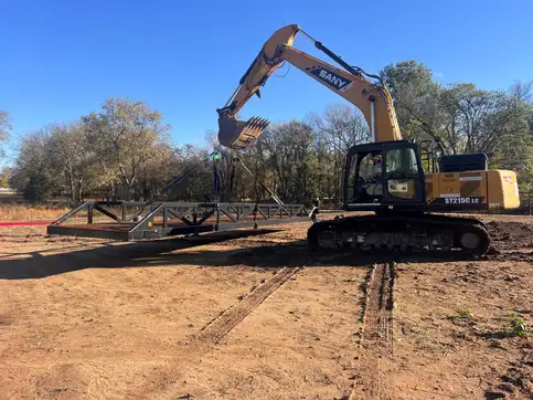 An excavator holding a steel truss bridge suspended on slings above graded ground, ready to place.