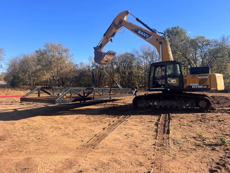 An excavator carrying an assembled steel truss bridge on slings.