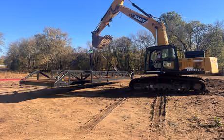 An excavator holding a steel truss bridge suspended on slings above graded ground, ready to place.