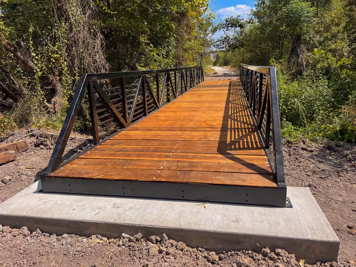 A finished steel trail bridge with timber decking and truss railing, seated on a poured concrete abutment, with a gravel path continuing beyond it.