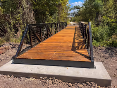 A finished steel trail bridge with timber decking and truss railing, seated on a poured concrete abutment, with a gravel path continuing beyond it.