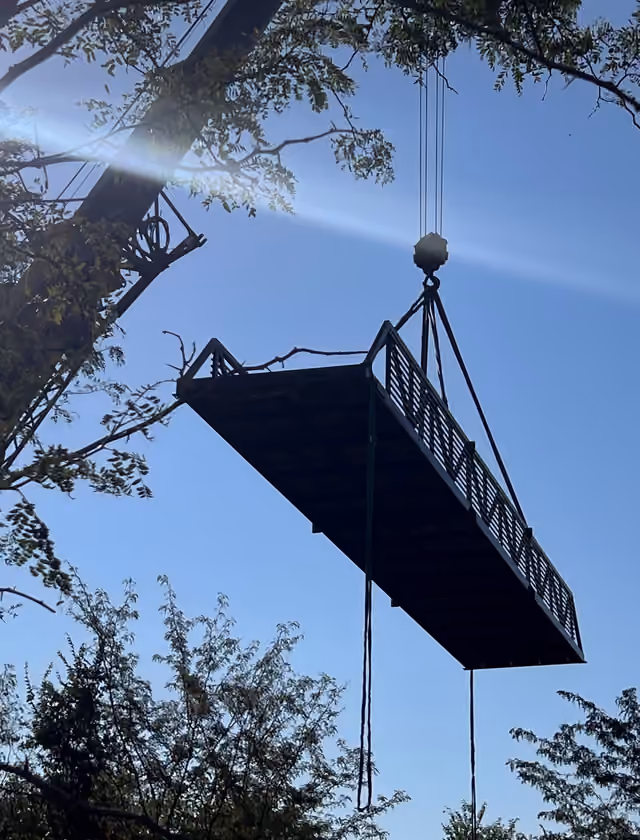 A steel bridge span hanging from a crane's slings high against a blue sky, in silhouette, with trees in the foreground and the sun flaring behind.