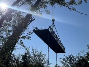 A steel bridge span hanging from a crane's slings high against a blue sky, in silhouette, with trees in the foreground and the sun flaring behind.