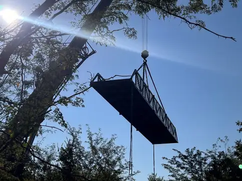 A steel bridge span hanging from a crane's slings high against a blue sky, in silhouette, with trees in the foreground and the sun flaring behind.