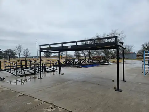 A bolt-together bridge standing on four adjustable steel legs on a concrete yard, with the shop and other bridges behind it.