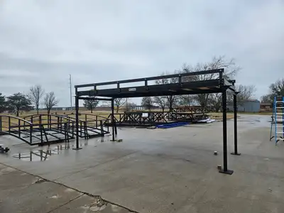 A bolt-together bridge standing on four adjustable steel legs on a concrete yard, with the shop and other bridges behind it.