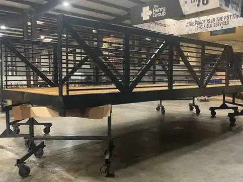 A black steel truss bridge with a rail of closely spaced horizontal bars and a timber deck, on wheeled stands in a workshop.
