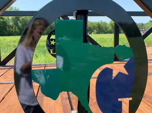 A circular steel medallion set into a bridge truss, cut with the outline of Texas and a letter G, painted green and blue against the black steel.