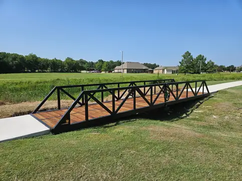 A black steel truss bridge carrying a concrete path across a grassed swale in a park, with houses beyond the treeline.
