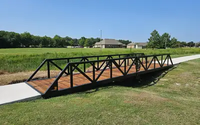A black steel truss bridge carrying a concrete path across a grassed swale in a park, with houses beyond the treeline.